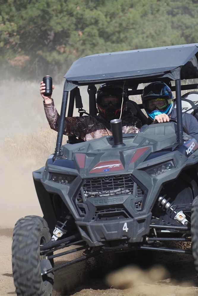 Two people in a side-by-side with the passenger raising their TwistLock Can Cooler up in the air while kicking up dust.